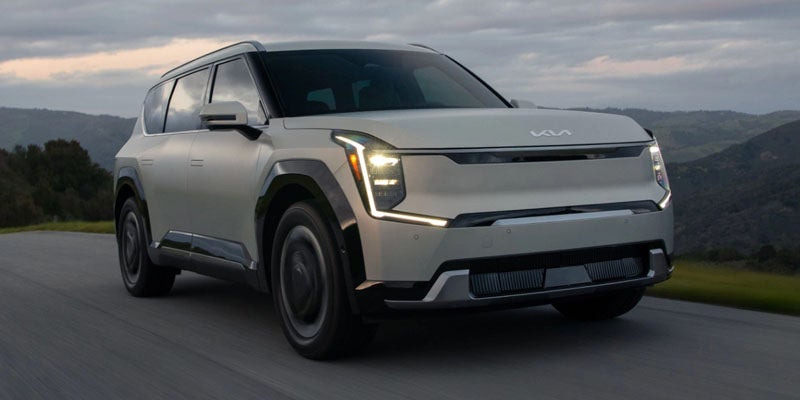 A silver Kia Sportage is driving on an open road with hills and cloudy skies