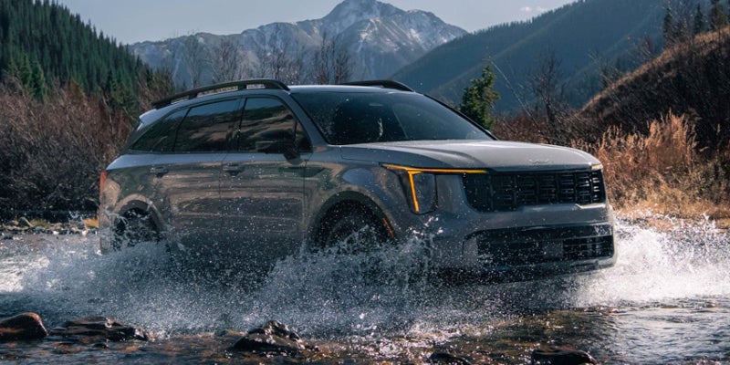 A dark-colored Kia drives through a shallow river