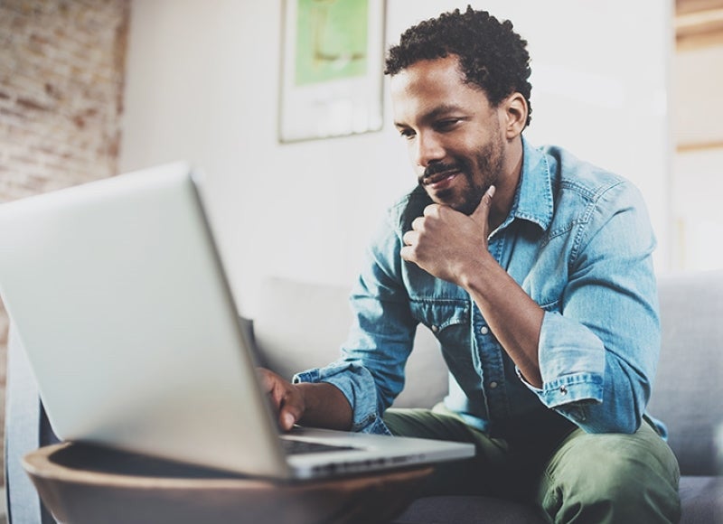 A man sitting on a sofa looking thoughtfully at his laptop screen.